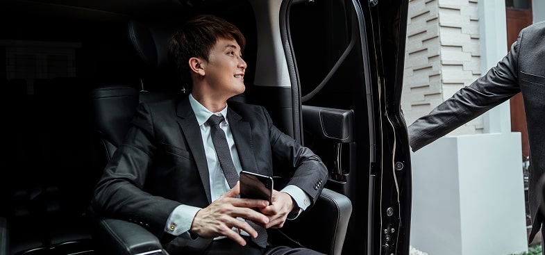 Business professional sitting inside a luxury black vehicle, smiling and holding a phone while chauffeur opens the door.