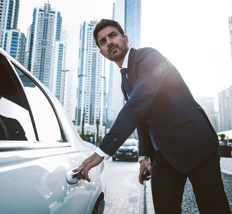 Business professional in a suit opening the door of a white luxury vehicle with a modern city skyline in the background.