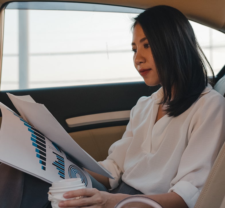 Businesswoman reviewing documents and holding a coffee cup while riding in the back seat of a luxury vehicle.